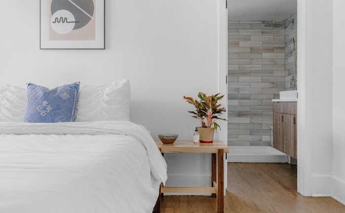 detalle de cama en blanco, con cojín azul, planta en la mesita y vista del baño