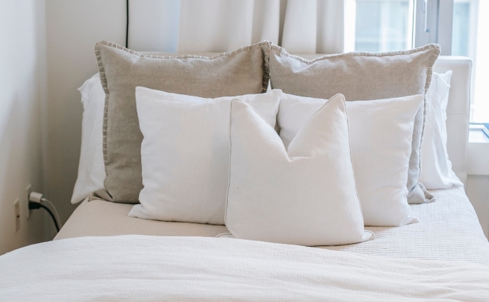 cama blanca con almohada y cojines blancos y dos cojines beige, con vista de parte de la ventana al fondo y dos enchufes