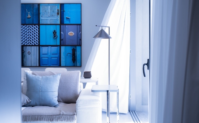 vista de salón en blancos, con cojines del sofá y cuadro al fondo en azules, mesa auxiliar blanca y lámpara de pie de metal