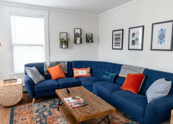 sofá en tonos azules con cojines naranjas en un salón con alfombra en los mismos tonos, mesa central de madera, plantas y cuadros en la pared